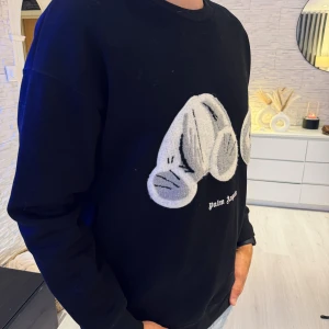 Palm Angels svart sweatshirt med nalle - Svart sweatshirt från Palm Angels med ikoniskt teddy-björn motiv i grått och vitt framtill. Tröjan har rund hals, långa ärmar och en relaxed passform. Perfekt statement-plagg för dig som gillar streetwear och coola detaljer. Passar bra som oversize modell.