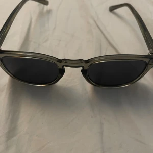 Svarta solglasögon med transparent båge - Snygga solglasögon med svarta linser och transparent gråbrun båge i klassisk rundad form. Bågen har en cool brygga och detaljer i metall vid gångjärnen. Perfekt accessoar för att höja din stil.