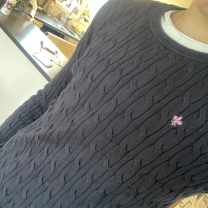 Mörkblå kabelstickad tröja Morris Lady - Kabelstickad mörkblå tröja från Morris Lady med rund halsringning och lång ärm. Tröjan har en rosa broderad lilja på bröstet och ribbade muddar vid ärmslut och nederkant. Perfekt för dig som gillar klassisk och stilren look.