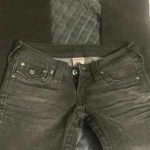 Svarta True Religion jeans, stl 24 - Snygga svarta jeans från True Religion med ikoniska bakfickor och silverfärgade knappar. Klassisk femficksmodell med markerade sömmar och logga på bakfickan. Jeansen är i stretchigt denim och har en smal passform som sitter riktigt bra.
