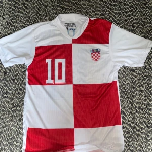 Kroatien Modric 10 fotbollströja M - Klassisk Kroatien fotbollströja med Modric och nummer 10 på ryggen. Tröjan har rödvita rutor, korta ärmar och är tillverkad i lätt, ventilerande polyester. Kroatiens landslagsmärke på bröstet och tryck i blått och rött. Perfekt för fotbollsfans!