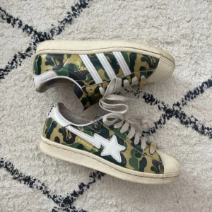 Adidas x Bape Superstar Camo Sneakers - BYD!!! Unika Adidas Superstar sneakers i samarbete med Bape. Skorna har klassiskt camouflagemönster i grönt, svart och beige, vita ränder och snörning samt Bape-stjärna på sidan. Guldiga detaljer på snörningen och loggor på häl och plös. Perfekta för dig som gillar streetwear och vill sticka ut.