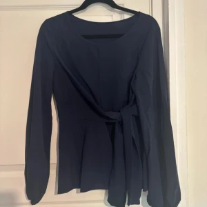 Mörkblå omlottblus med knytband - Snygg mörkblå blus med långa ärmar och rund halsringning. Toppen har ett omlottliv med knytband i midjan som ger en schysst siluett. Ärmarna är lite puffiga med resår vid handlederna. Perfekt för dig som gillar stilrena och enkla plagg med en twist.