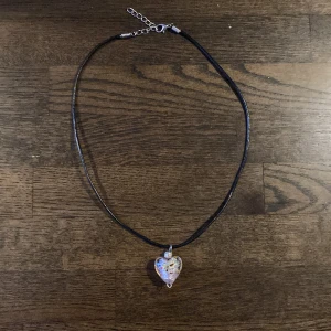 Halsband med hjärthänge i glas - Svart halsband med justerbart spänne och ett rosa hjärthänge i glas. Hänget har färgglada inslag i rött, gult, grönt och blått, vilket ger en unik och däegglad detalj. Perfekt accessoar för att lyfta din outfit med lite färg och personlighet.