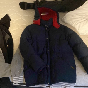 Mörkblå dunjacka från Polo Ralph Lauren - Säljer en mörkblå dunjacka från Polo Ralph Lauren med röd insida på huvan och kragen. Jackan har en klassisk pufferstil, dragkedja och tryckknappar framtill samt två stora fickor. Perfekt för kalla vinterdagar och riktigt snygg streetstil.