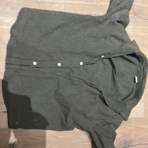 Mörkgrön linneskjorta med knappar - Säljer en mörkgrön skjorta i linne med klassisk krage och vita knappar framtill. Skjortan har lång ärm och en avslappnad passform, perfekt att styla med jeans eller shorts. Enkel och stilren design som funkar året runt.