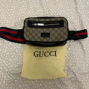 Gucci axelväska med GG-mönster - Snygg axelväska från Gucci med klassiskt GG-mönster i beige och brunt. Väskan har svarta skinn-detaljer, silverfärgad dragkedja och bred axelrem i grönt och rött. Perfekt för dig som vill ha en ikonisk och trendig accessoar.