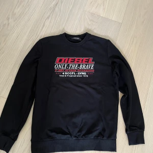 Svart sweatshirt från Diesel - Svart sweatshirt från Diesel med tryck i rött och vitt framtill. Klassisk rund halsringning och långa ärmar med muddar. Snygg streetstil med texten 'ONLY THE BRAVE' och detaljerade tryck. Perfekt för dig som gillar en clean och cool look.