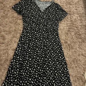 Svart blommig klänning  - Svart klänning med vita blommor. Korsad omlott och knä längd! Aldrig använd.
