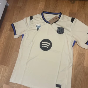 Beige FC Barcelona Nike fotbollströja - FC Barcelona bortatröja från Nike i beige med svarta och blå detaljer. Tröjan har korta ärmar, svart klubbmärke, Spotify-logga och UNHCR-tryck på ryggen. Tillverkad i lätt Dri-FIT-material som andas. Perfekt för dig som älskar Barca och snygg design.