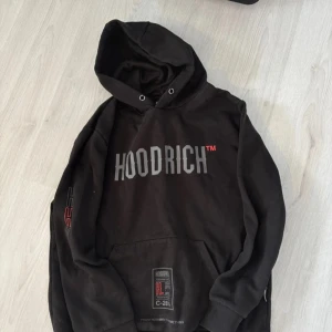 Svart hoodie från Hoodrich XL - Svart hoodie från Hoodrich i storlek XL med stor logga på bröstet och tryck på ryggen. Tröjan har huva, känguruficka och coola detaljer i grått och rött. Perfekt för streetwear-stilen och chill dagar. Materialet är mjukt och skönt, troligen bomull och polyester.
