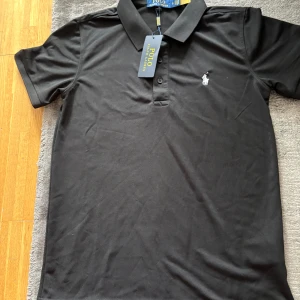 Svart Polo Ralph Lauren piké S - Svart piké tröja från Polo Ralph Lauren med klassisk pikékrage och två knappar framtill. Ikonisk broderad logga i vitt på bröstet. Tillverkad i mjuk bomull som känns skön mot huden. Perfekt för dig som gillar stilrena och tidlösa plagg.