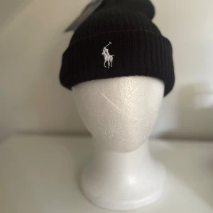 Svart ribbad mössa Polo Ralph Lauren - Svart ribbstickad mössa från Polo Ralph Lauren med klassisk vit broderad logga framtill. Tjockt uppvik och stilren design som passar perfekt till kalla dagar. Tillverkad i mjukt material för extra komfort.