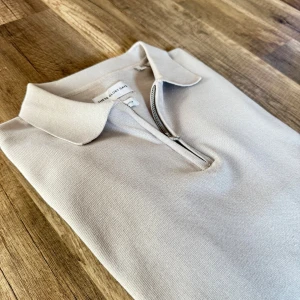 Beige halvzip-tröja polo/ pike från Three Glory Days - Stilren beige långärmad tröja från Three Glory Days med klassisk krage och halv dragkedja framtill. Tröjan är i mjukt material och har ribbade muddar vid ärmslut och nederkant. Perfekt för en clean och avslappnad look.