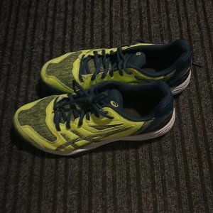 Asics gula och blå tennisskor/padelskor  - Säljer ett par Asics tennisskor i en fräsch gul och mörkblå färgkombination. Skorna har snörning, platt sula med fiskbensmönster och är tillverkade i syntetmaterial och mesh för bra andningsförmåga. som nya 