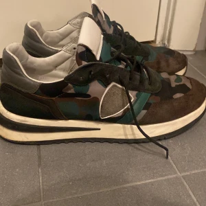 Philippe Model sneakers camouflage - Snygga sneakers från Philippe Model med camouflage-mönster i grönt, svart och grått. Skorna har detaljer i mocka och skinn, vit plös och svart/vit sula. Klassisk snörning och rund tå ger en sportig vibe. Perfekta för dig som gillar streetstyle och unika detaljer.