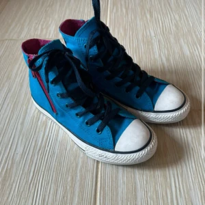 Blå Converse All Star high-tops - Säljer ett par blå Converse All Star high-tops med svarta skosnören och vit tå. Skorna har rosa detaljer på insidan och dragkedja på sidan. Klassisk rund tå och vit sula. Tillverkade i textil med Converse-logga på sidan.