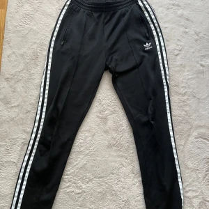 Svarta Adidas mjukisbyxor med ränder - Svarta mjukisbyxor från Adidas med klassiska vita ränder längs benen och broderad logga på höften. Byxorna har resår i midjan, dragkedjefickor och smal passform. Perfekta för chill eller träning.