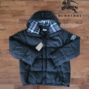 Burberry Jacka - Svart dunjacka från Burberry med klassisk Säljer en elegant och mångsidig Burberry-jacka i svart, i nyskick. Dunfodrad för värme och komfort med det klassiska rutmönstret i huvan som ger en exklusiv touch. Praktiska dragkedjefickor, avtagbar huva och diskret logopatch på ärmen. Sitter som (M/L). Jackan är i princip som ny och utan defekter. En lyxig kombination av stil och funktion – perfekt för både vardag och tillfällen då du vill klä upp dig.  📦Snabb leverans - skickas samma dag📦 