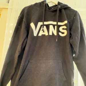 Svart hoodie från Vans med logga - Svart hoodie från Vans i storlek small med stor vit Vans-logga framtill. Klassisk känguruficka, huva med snörning och långa ärmar. Perfekt för en chill och avslappnad stil. Solen ligger på därav den blekta effekten hamnade på magen.