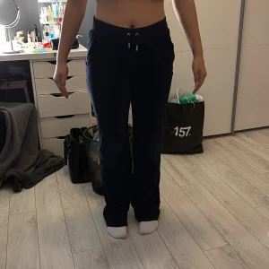 Mörkblå mjukisbyxor Juicy Couture S - Mörkblå mjukisbyxor från Juicy Couture med snörning i midjan och raka ben. Byxorna har fickor fram och bak med knappdetaljer. Perfekta för en chill och avslappnad stil.