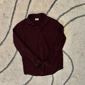 Vinröd skjorta från Weekday - Vinröd skjorta från Weekday i mjuk bomull. Klassisk krage, bröstficka och knäppning framtill. Långärmad och passar perfekt till lager-på-lager-stil. Enkel och stilren design som funkar till många outfits.