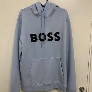 Ljusblå hoodie från BOSS - Snygg ljusblå hoodie från BOSS med stor svart logga framtill. Tröjan har huva med dragsko, känguruficka och långa ärmar. Tillverkad i mjukt syntetmaterial med meshfoder i huvan för extra komfort. Perfekt för en chill och stilren look.