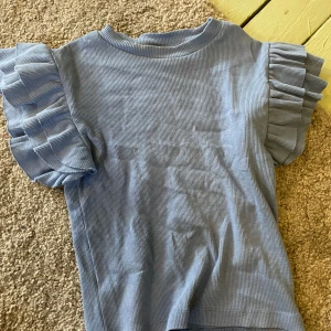 Blå ribbad topp med volangärmar Gina Tricot - Supersöt ljusblå ribbad topp från Gina Tricot med korta volangärmar som ger en extra touch. Toppen har rund halsringning och är gjord i mjuk bomull, perfekt för en chill och trendig look. Väldigt söt i storlek 140,