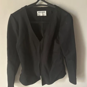 Svart kostym kofta från AWARE med v-ringning - Svart kofta från AWARE med djup v-ringning och långa ärmar. Stängs framtill med diskreta hyskor. Enkel och stilren design som passar perfekt till lager-på-lager. Mjukt material som känns skönt mot huden.