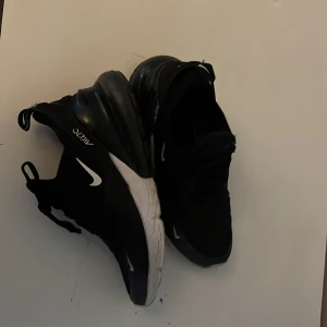 Nike Air Max 270 svarta sneakers - Svarta Nike Air Max 270 sneakers med vit sula och diskreta röda detaljer undertill. Skorna har en luftig mesh-överdel, tydlig Air Max-dämpning i hälen och klassisk snörning. Swoosh-logga på sidorna och 270-tryck. Perfekta för dig som gillar sportig och modern stil. Säljer på grund av förstora