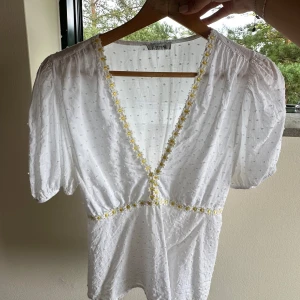 Vit blus med broderade blommor - Superfin vit blus med v-ringning och kort puffärm. Blusen har små broderade prickar i tyget och söta gula blommor längs urringningen och midjan. 
