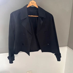 Svart kort trenchcoat från 157 - Kort svart trenchcoat från 157 med dubbelknäppning och bred krage. Jackan har långa ärmar med knappar vid ärmsluten. 