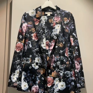 Blommig kostym från H&M, strl 34 - Säljer en kostym från H&M, st 34 på kavajen och 36 i byxorna. Kostymen har svart botten med färgglatt blommigt mönster i rosa, blått, vitt och gult. Kavajen har klassisk passform med knappar och fickor, och byxorna har matchande blommigt tryck och knappstängning. 