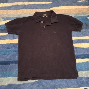 Mörkblå pikétröja från Giorgio Armani junior - Klassisk mörkblå pikétröja från Giorgio Armani junior med korta ärmar och krage med två knappar. Tröjan är i bomull och har en diskret broderad logga på bröstet. Perfekt för en stilren och avslappnad look. Säljer då den är för liten för mig. Passar folk som är 140-154 isch. Ny pris ca 1500-2000kr Pris kan diskuteras.