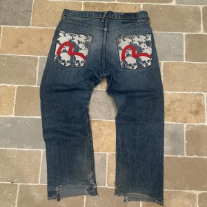 Evisu jeans - Unika Evisu jeans i mörkblå denim med raka ben och råa, slitna benslut. Bakfickorna har ett grått/vitt dödskallemönster med en fet röd broderad båge. Klassisk femficksmodell och knappgylf. Perfekta för dig som vill sticka ut.