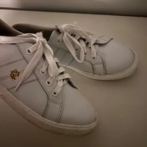 Vita sneakers från Ralph Lauren i storlek 39 - Snygga vita sneakers från Ralph Lauren med klassisk design och guldfärgad RL-detalj på sidan. Skorna har snörning, rund tå och är gjorda i skinn med vita sömmar och vit sula. Perfekta för en clean och stilren look.