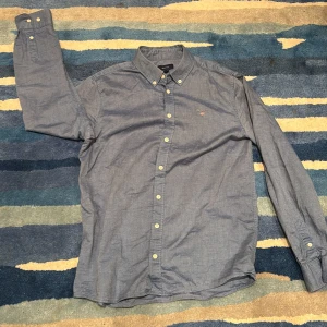 Blå skjorta från GANT med knappar - Snygg blå skjorta från GANT med klassisk button down-krage och vita knappar. Skjortan har broderad GANT-logga på bröstet. Pris kan diskuteras