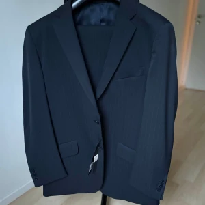 Mörkblå randig kostym – Rice, Made in Italy (stl 54) - Elegant mörkblå kostym med subtila ränder från det italienska kvalitetsmärket Rice. Klassisk och tidlös design som passar perfekt för både affärssammanhang och festligare tillfällen. Tillverkad i fin ullblandning med fokus på passform och komfort.  Storlek: 54  Färg: Mörkblå med diskreta ränder  Modell: R206  Material: 100% virgin wool  Nypris: ca 4685 kr  Skick: Helt ny med tags kvar