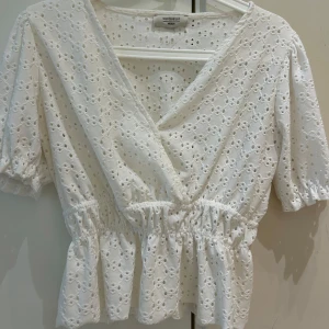 Vit broderad blus från Chiquelle - Superfin vit blus från Chiquelle med broderade detaljer och omlott framtill. Blusen har korta puffärmar med resår och markerad midja med volangkant. 