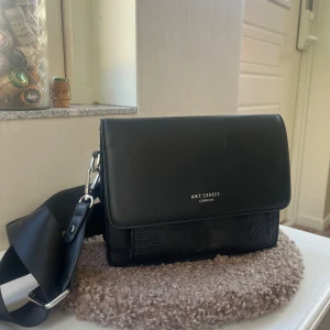 Svart axelväska från Art Street London - Snygg svart axelväska med bred justerbar rem och croco-detaljer på framsidan. Väskan har stilren design med silverfärgade metalldetaljer och logga framtill. Perfekt storlek för mobil, plånbok och andra essentials. Materialet är skinnimitation.