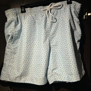 Blåvita mönstrade badbyxor - Shorts i ljusblått och vitt med geometriskt mönster. Materialet känns lätt och luftigt, perfekt för varma dagar. Fickor på sidorna och bak för extra funktion.