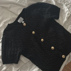 Svart stickad kofta från H&M med guldknappar - Svart stickad kofta från H&M med korta ärmar och snygga guldknappar framtill. Koftan har ett flätstickat mönster och ribbade muddar vid hals och ärmslut. Perfekt för dig som vill ha en stilren men ändå trendig look.