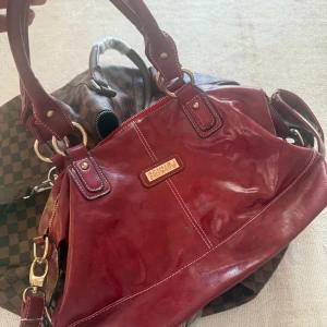 Vinröd handväska från Beinipj - Vinröd handväska i skinn från Beinipj med snygga guldfärgade metalldetaljer och vita kontrastsömmar. Väskan har både handtag och axelrem, dragkedja upptill samt flera fack inuti och ett ytterfack med dragkedja. Perfekt storlek för allt du behöver ha med dig.