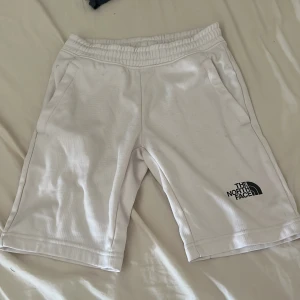 Vita shorts från The North Face - Snygga vita shorts från The North Face med elastisk midja och fickor på sidorna. Loggan syns både fram och längs benet i svart. Perfekta för chill dagar eller träning, tillverkade i mjuk bomull för skön känsla. Passformen är normal och längden går strax ovanför knäna.