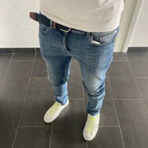 Blå jeans från Tiger Of Sweden - Säljer ett par snygga blå jeans från Tiger Of Sweden modell Leon. Jeansen har klassisk femficksdesign, normal passform och raka ben. De är tillverkade i bomull och har en schysst tvättad look med ljusare partier på låren. Perfekta för dig som gillar stilrena och bekväma jeans.