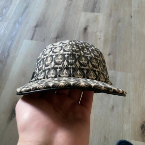 Beige och svart keps från Ferragamo - Snygg keps från Salvatore Ferragamo med beige och svart mönster över hela kepsen. Justerbar rem baktill med metallspänne. Tillverkad i bomull och har en cool, grafisk design som sticker ut. Perfekt för dig som vill ha något unikt och stilrent.