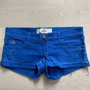 Blå jeansshorts från Hollister - Snygga blå jeansshorts från Hollister med låg midja och slitna detaljer. De har klassiska fem fickor, uppvikta benslut med rå kant och logga på fickan. Perfekta för varma dagar och har en cool, avslappnad vibe.