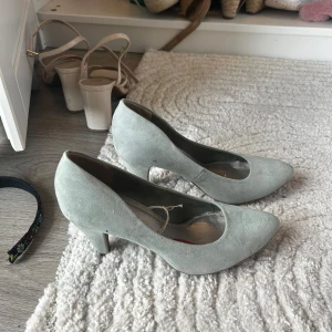 Ljusgrå mockapumps från Marco Tozzi - Snygga ljusgrå pumps från Marco Tozzi i mjuk mocka. Skorna har spetsig tå och en smal klack som ger en elegant look. Perfekta för dig som vill ha en stilren och klassisk sko med modern touch.