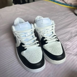 Nike Jordan 1 Low svart/vit sneakers - Nike Jordan 1 Low sneakers i svart och vitt med klassisk Swoosh på sidan. Skorna har snörning, perforerad tå och platt sula. Ovandelen är i skinn och mesh, vilket ger både stil och komfort. Perfekt för dig som gillar clean och tidlös streetstil.