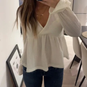 Vit omlottblus med v-ringning - Supersöt vit blus med omlott och v-ringning. Lätt och luftig med långärmade ärmar och volangkant vid ärmslut. Perfekt till jeans eller kjol för en fräsch look. Materialet känns mjukt och har en lätt struktur.